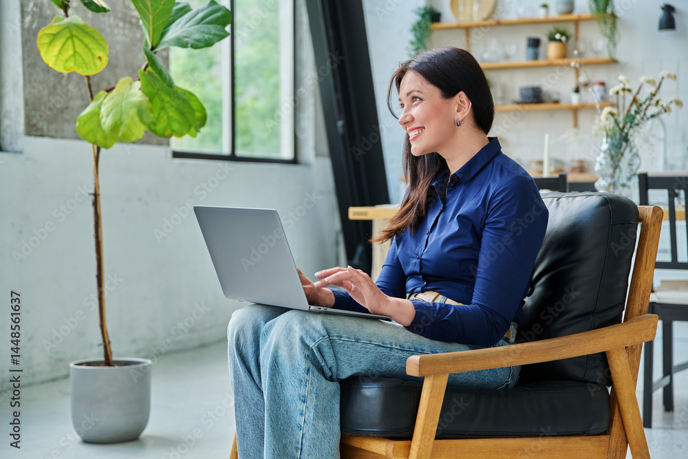 Young smiling woman typing on laptop sitting on chair at home