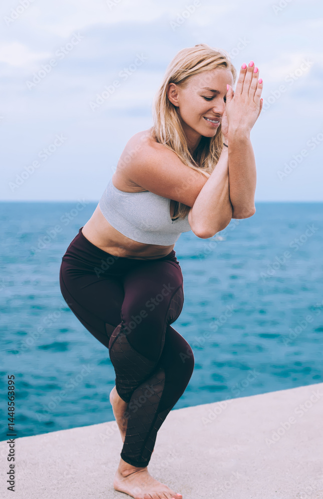 Healthy lifestyle exercise concept - barefoot Caucasian woman practices balance yoga asana garudasana near ocean, sportive girl in tracksuit doing eagle pose for recreating soul and full minds