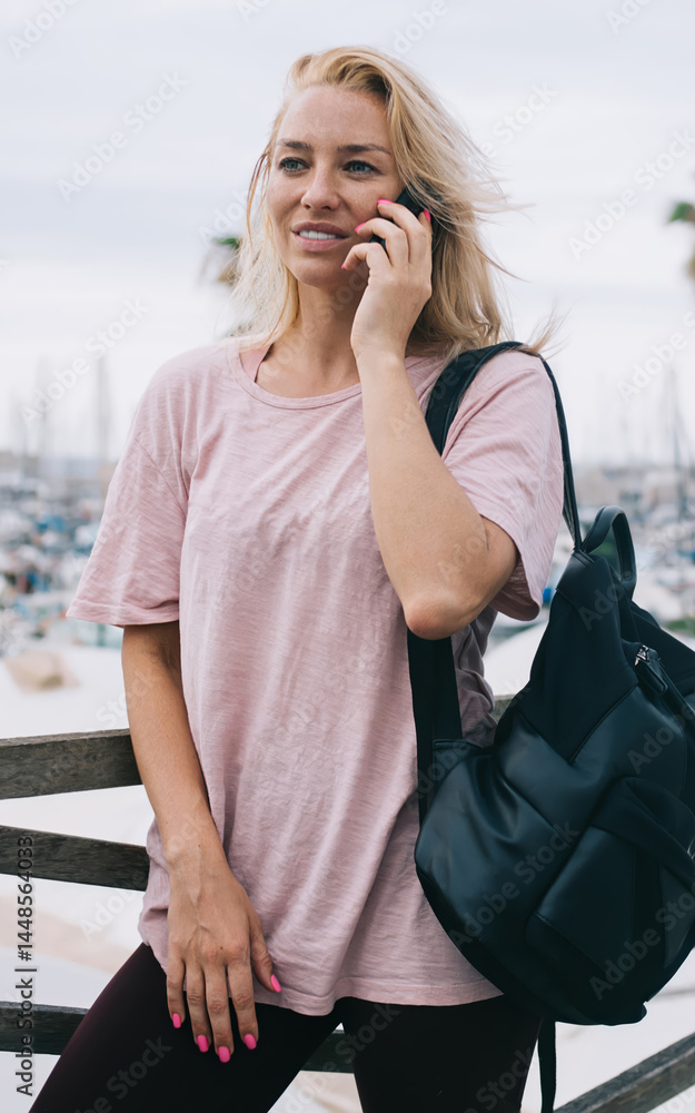 Attractive female with touristic backpack receiving cellphone call for communicate in roaming, casual dressed travel girl phoning via mobile application spending time for international conversation