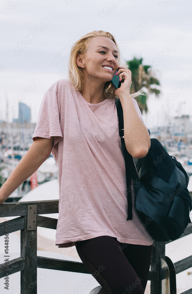 Happy young woman connecting to roaming 4g for making friendly international conversation during solo travelling, funny female user with backpack enjoying phone conversation at urban setting