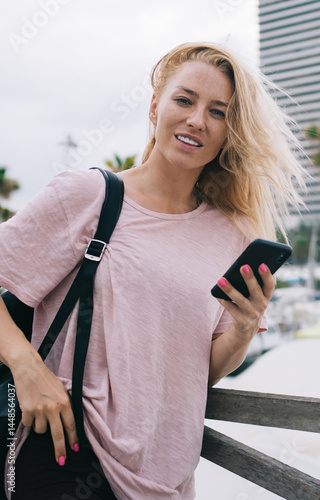 Obraz na płótnie Half length portrait of charming Caucasian millennial user holding modern cellphone device in hand, beautiful female traveller with touristic backpack connecting to 4g wireless on smartphone gadget w salonie