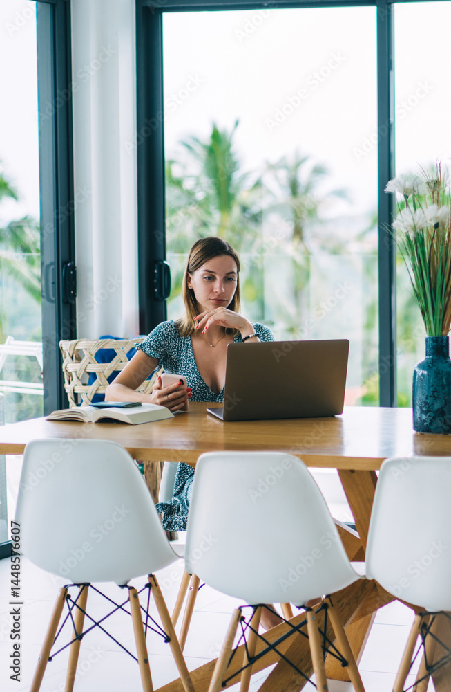 Focused woman watching video on netbook while using smartphone in modern living room