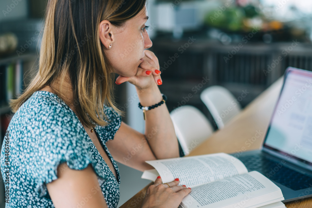 Concentrated young woman using laptop and reading book while working in cozy cafe