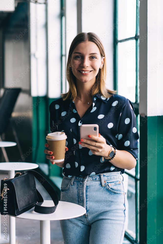 Cheerful female with coffee and smartphone smiling at camera