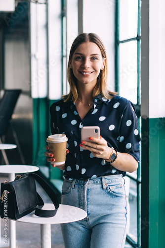 Obraz na płótnie Cheerful female with coffee and smartphone smiling at camera w salonie