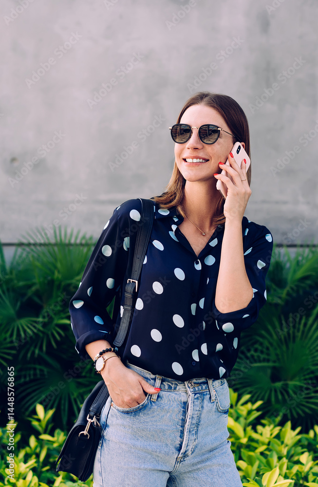 Positive young woman talking on smartphone while standing on background of stone wall and tropical plants