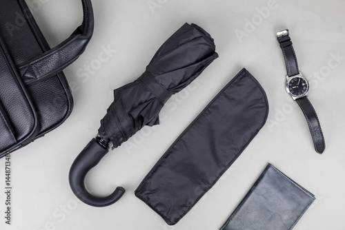 A black umbrella in a case lies on a table next to a man's watch and a leather bag
