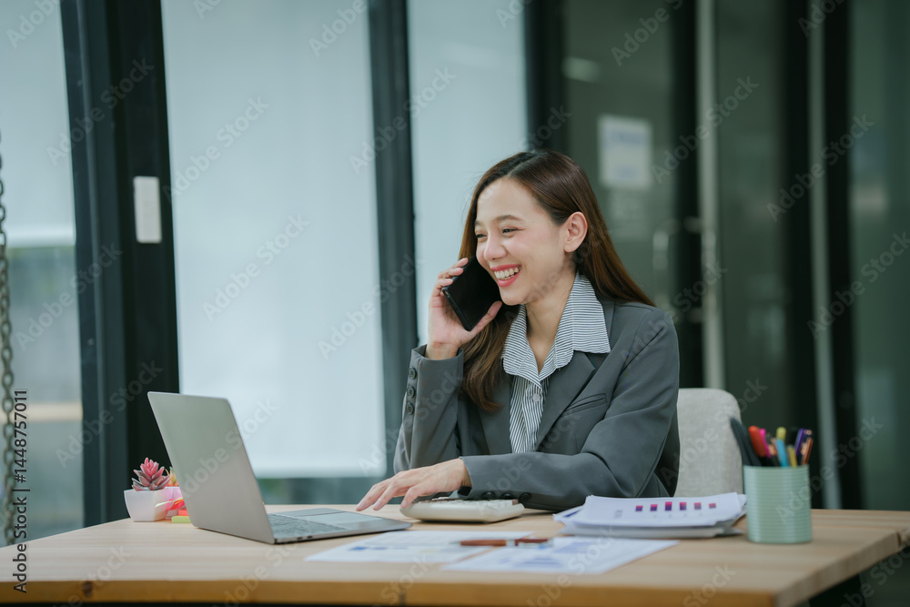 Smiling asian businesswoman talking on phone and using laptop while working at office desk Smiling asian businesswoman talking on phone and using laptop while working at office desk
