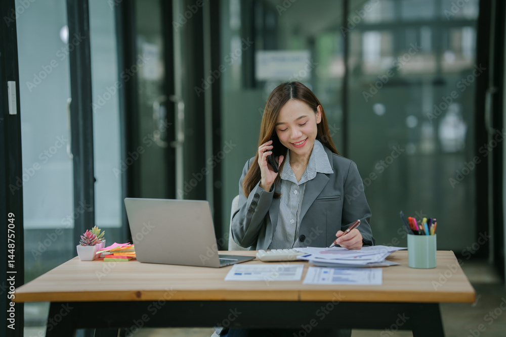 Smiling asian businesswoman talking on phone and using laptop while working at office desk Smiling asian businesswoman talking on phone and using laptop while working at office desk