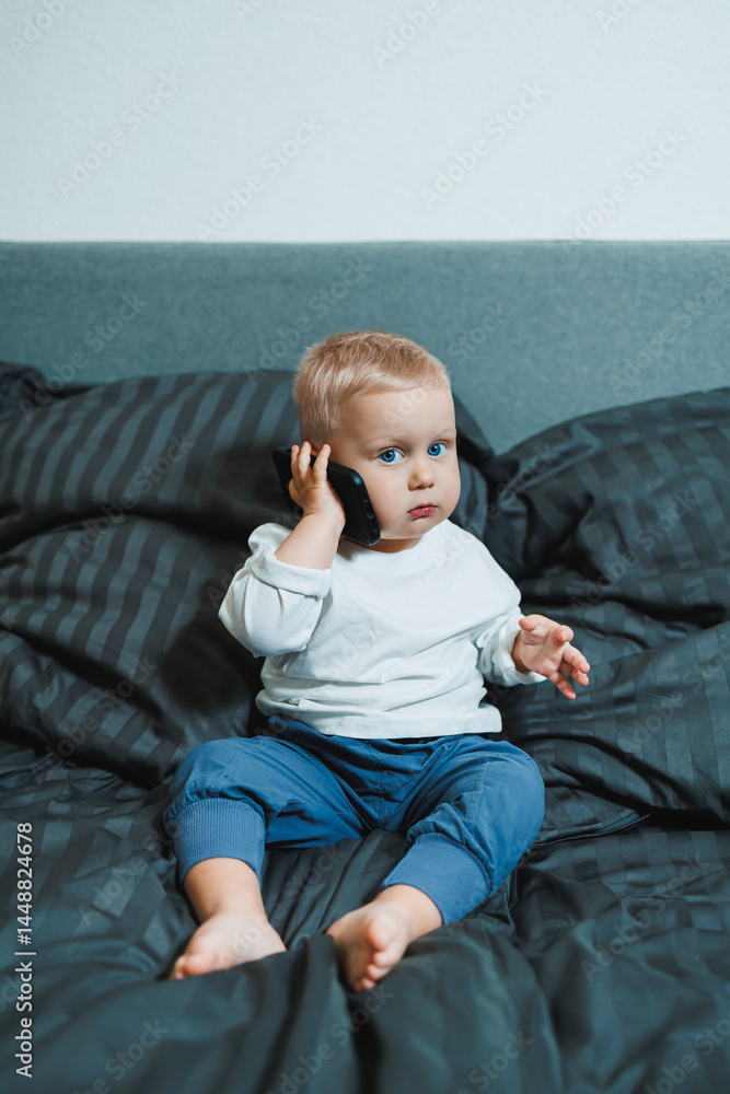 Two-year-old little boy in a white T-shirt sits on the bed with a phone in his hands. Digitalization of modern little children. Toddler boy studies mobile phone.
