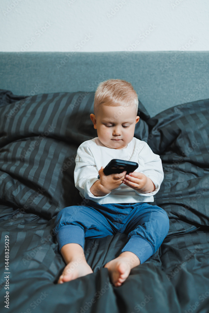 Cheerful toddler boy sitting on bed and playing on smartphone during day at home. Close up of child sitting on bed holding smartphone at home. Smiling child with mobile.