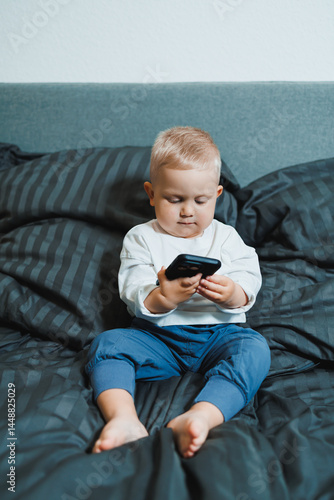 Obraz na płótnie Cheerful toddler boy sitting on bed and playing on smartphone during day at home. Close up of child sitting on bed holding smartphone at home. Smiling child with mobile. w salonie