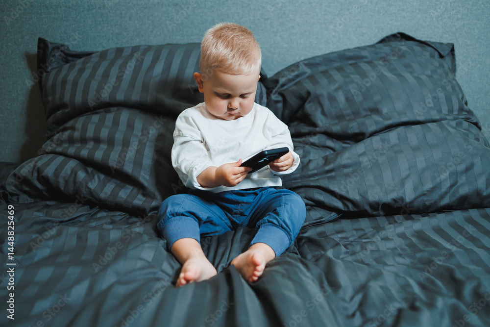 Two-year-old little boy in a white T-shirt sits on the bed with a phone in his hands. Digitalization of modern little children. Toddler boy studies mobile phone.