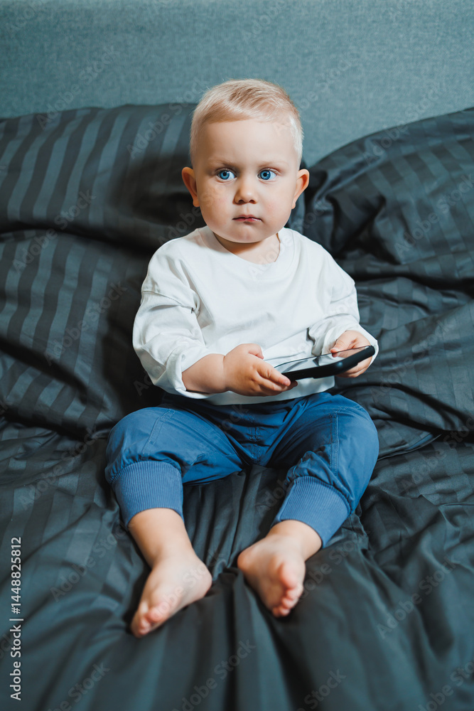 Cheerful toddler boy sitting on bed and playing on smartphone during day at home. Close up of child sitting on bed holding smartphone at home. Smiling child with mobile.