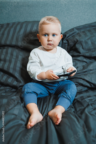 Obraz na płótnie Cheerful toddler boy sitting on bed and playing on smartphone during day at home. Close up of child sitting on bed holding smartphone at home. Smiling child with mobile. w salonie