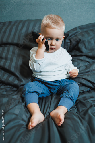 Obraz na płótnie Two-year-old little boy in a white T-shirt sits on the bed with a phone in his hands. Digitalization of modern little children. Toddler boy studies mobile phone. w salonie