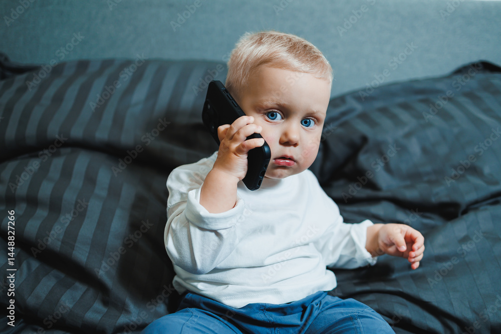 Cheerful toddler boy sitting on bed and playing on smartphone during day at home. Close up of child sitting on bed holding smartphone at home. Smiling child with mobile.
