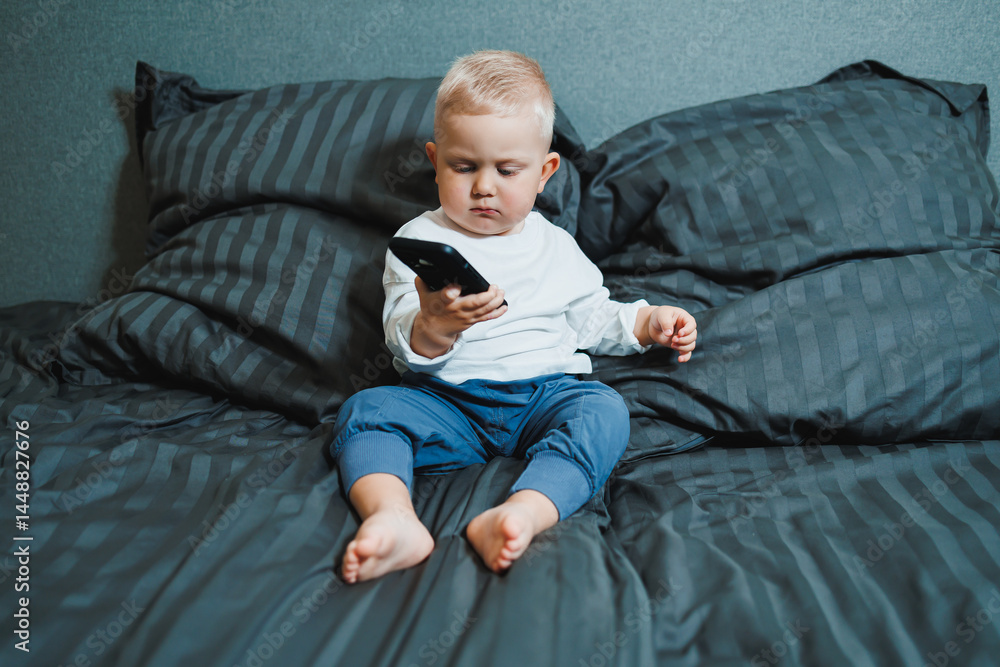 Cute little boy 2 years old using smartphone, looking at screen, child holding phone in hands, sitting on sofa at home alone, playing game on mobile device, watching cartoons online