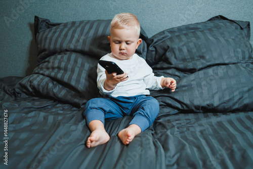 Obraz na płótnie Cute little boy 2 years old using smartphone, looking at screen, child holding phone in hands, sitting on sofa at home alone, playing game on mobile device, watching cartoons online w salonie