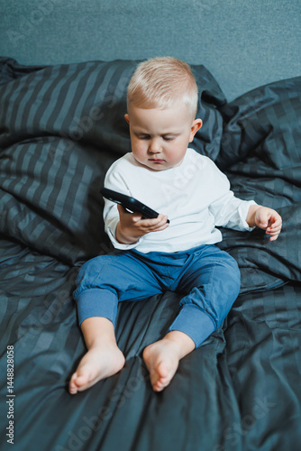 Boy 2 years old with a smartphone. Toddler boy sitting in bed and playing with a mobile phone. Calling mom. Cute boy holding a mobile phone in his hands and looking intently at the screen.