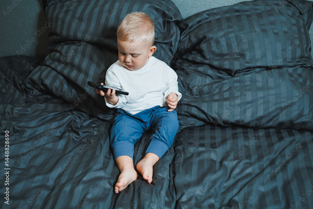 Cute little boy 2 years old using smartphone, looking at screen, child holding phone in hands, sitting on sofa at home alone, playing game on mobile device, watching cartoons online