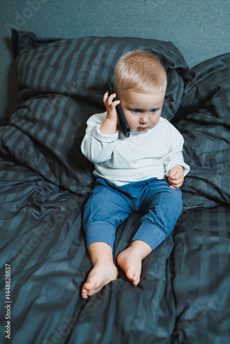 Obraz na płótnie Boy 2 years old with a smartphone. Toddler boy sitting in bed and playing with a mobile phone. Calling mom. Cute boy holding a mobile phone in his hands and looking intently at the screen. w salonie