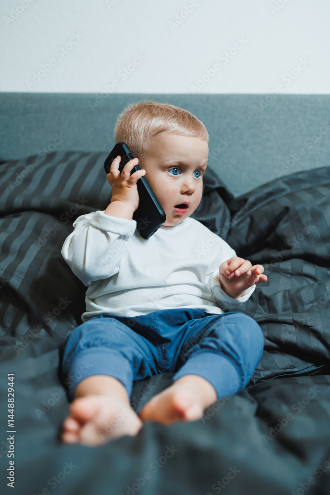 Toddler boy sitting on bed, deeply focused on playing on mobile phone. He is wearing a t-shirt and playing a game on his phone. Modern digitalization of children. Toddler with mobile phone