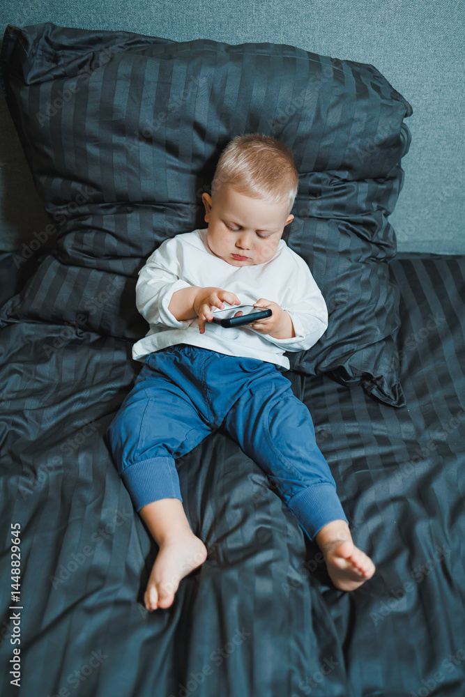 Toddler boy sitting on bed, deeply focused on playing on mobile phone. He is wearing a t-shirt and playing a game on his phone. Modern digitalization of children. Toddler with mobile phone
