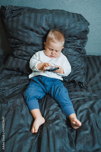 Obraz na płótnie Toddler boy sitting on bed, deeply focused on playing on mobile phone. He is wearing a t-shirt and playing a game on his phone. Modern digitalization of children. Toddler with mobile phone w salonie