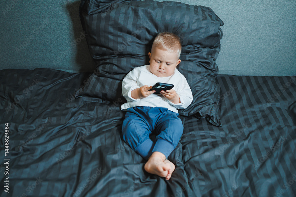 Toddler boy sitting on bed, deeply focused on playing on mobile phone. He is wearing a t-shirt and playing a game on his phone. Modern digitalization of children. Toddler with mobile phone