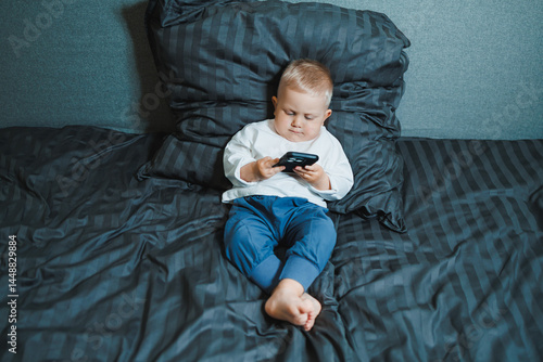 Obraz na płótnie Toddler boy sitting on bed, deeply focused on playing on mobile phone. He is wearing a t-shirt and playing a game on his phone. Modern digitalization of children. Toddler with mobile phone w salonie