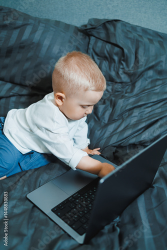 Little boy toddler using laptop watching video at home, early education and learning. Little child looking at laptop screen and listening to music while sitting on bed.