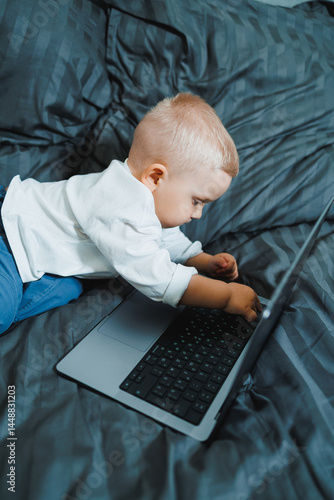 Obraz na płótnie Cute boy 2 years old sits in bed and presses keys on laptop. Child sits and watches video on laptop. Modern development and digitalization of children. w salonie