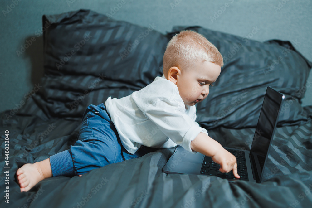 Cute boy 2 years old sits in bed and presses keys on laptop. Child sits and watches video on laptop. Modern development and digitalization of children.