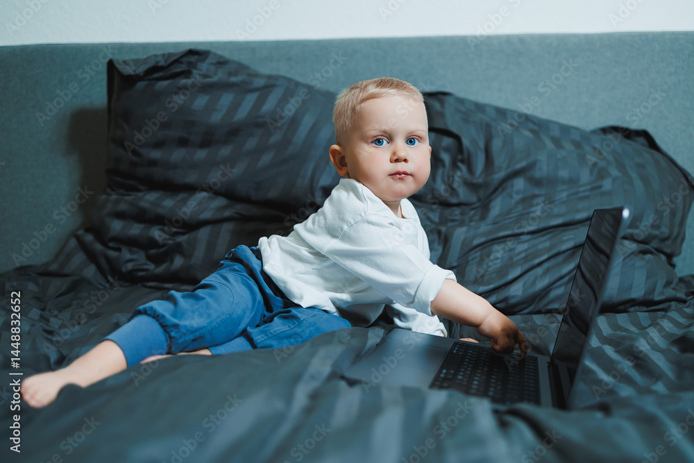 Cute boy 2 years old sits in bed and presses keys on laptop. Child sits and watches video on laptop. Modern development and digitalization of children.