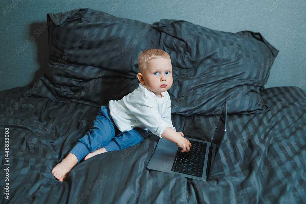 Little boy toddler using laptop watching video at home, early education and learning. Little child looking at laptop screen and listening to music while sitting on bed.