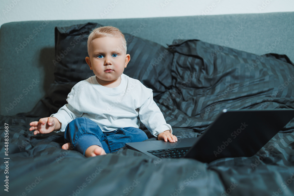 Stylish blond toddler boy sitting on bed and playing with laptop at home in bedroom. Child with tablet computer watching cartoons. White cozy interior in bedroom.