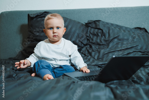 Obraz na płótnie Stylish blond toddler boy sitting on bed and playing with laptop at home in bedroom. Child with tablet computer watching cartoons. White cozy interior in bedroom. w salonie