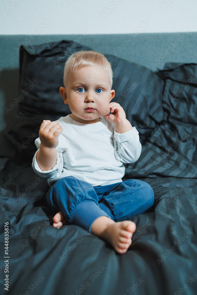 Two-year-old blue-eyed boy in a white long-sleeved shirt and pants sits in bed. Toddler boy in home clothes sits in the bedroom on the bed.