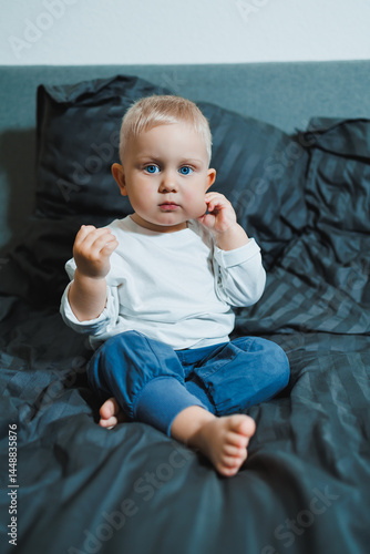 Obraz na płótnie Two-year-old blue-eyed boy in a white long-sleeved shirt and pants sits in bed. Toddler boy in home clothes sits in the bedroom on the bed. w salonie