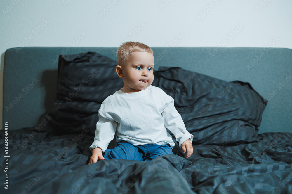 Child in bed. Cute toddler boy in white blouse and pants laughing while sitting in bed. 2 year old boy playing while sitting in bed. Home clothes for boys.
