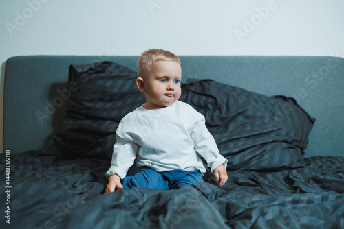 Obraz na płótnie Child in bed. Cute toddler boy in white blouse and pants laughing while sitting in bed. 2 year old boy playing while sitting in bed. Home clothes for boys. w salonie