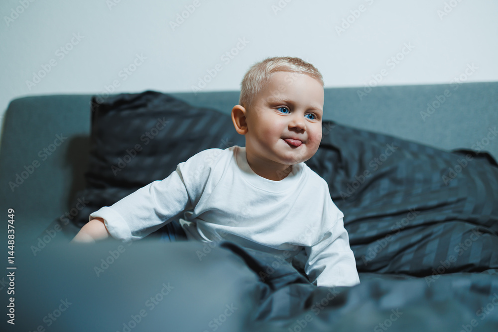 Cute toddler boy in white shirt and pants laughing while sitting in bed. 2 year old boy playing while sitting in bed. Home clothes for boys.