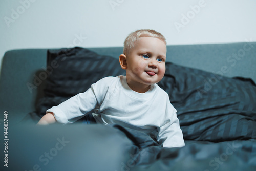 Cute toddler boy in white shirt and pants laughing while sitting in bed. 2 year old boy playing while sitting in bed. Home clothes for boys.