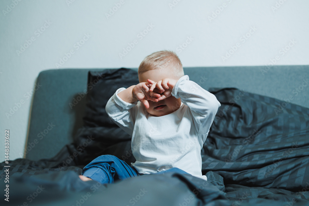 Two-year-old blue-eyed boy in a white long-sleeved shirt and pants sits in bed. Toddler boy in home clothes sits in the bedroom on the bed.