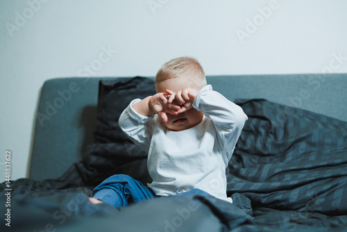 Two-year-old blue-eyed boy in a white long-sleeved shirt and pants sits in bed. Toddler boy in home clothes sits in the bedroom on the bed.