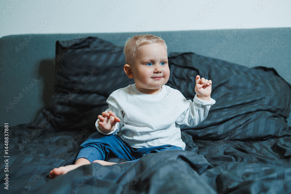 Child in bed. Cute toddler boy in white blouse and pants laughing while sitting in bed. 2 year old boy playing while sitting in bed. Home clothes for boys.