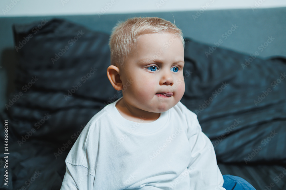 Child in bed. Cute toddler boy in white blouse and pants laughing while sitting in bed. 2 year old boy playing while sitting in bed. Home clothes for boys.