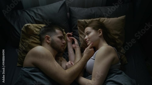 Top view of young man and woman in underwear lying in bed at home, facing each other, enjoying bond and connection. Gentle touches, subtle smiles, soft look in eyes, love in air, copy space
