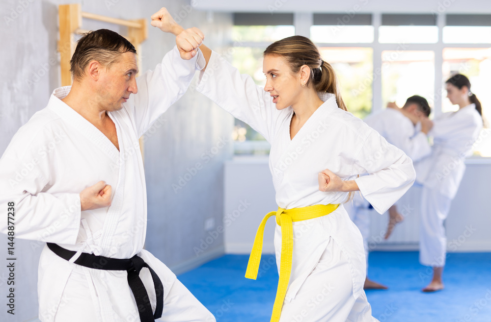 Fototapeta premium Adult man and young woman in kimono practicing karate techniques in group in gym..